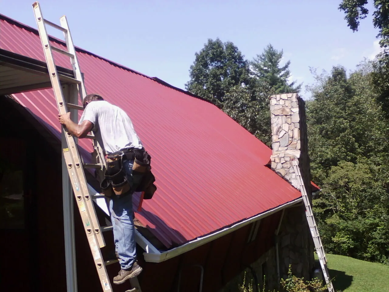 Roofer on a ladder beside a red metal roof and stone chimney — Soffit & Fascia Repair by Roofing Brothers in Woolstock